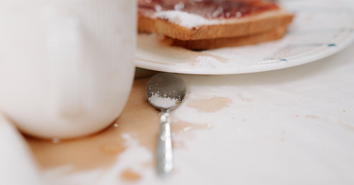 Metal spoon on a messy white tabletop beside a mug and a plate with toast.