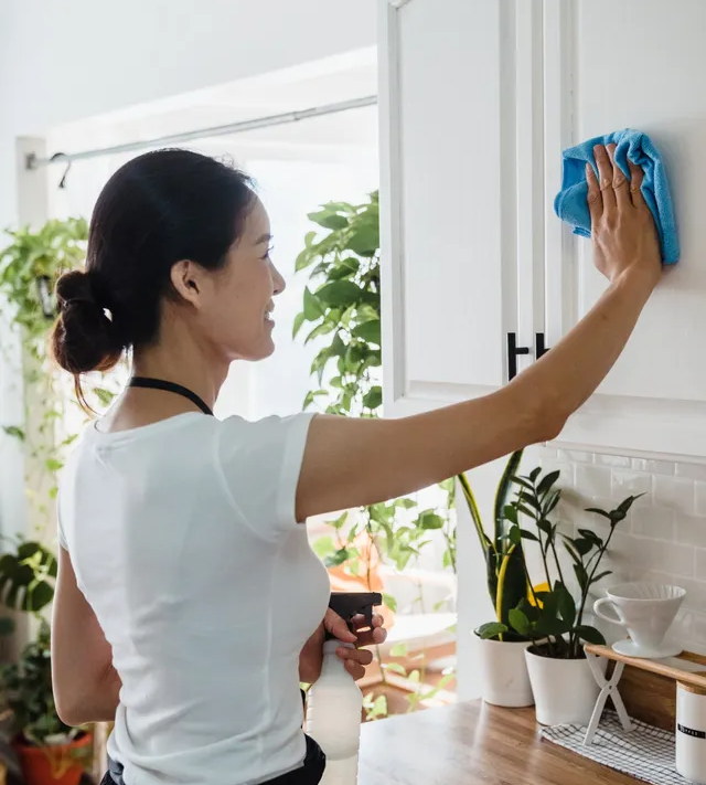 A person in a white shirt cleans a white kitchen cabinet with a blue cloth while holding a spray bottle.