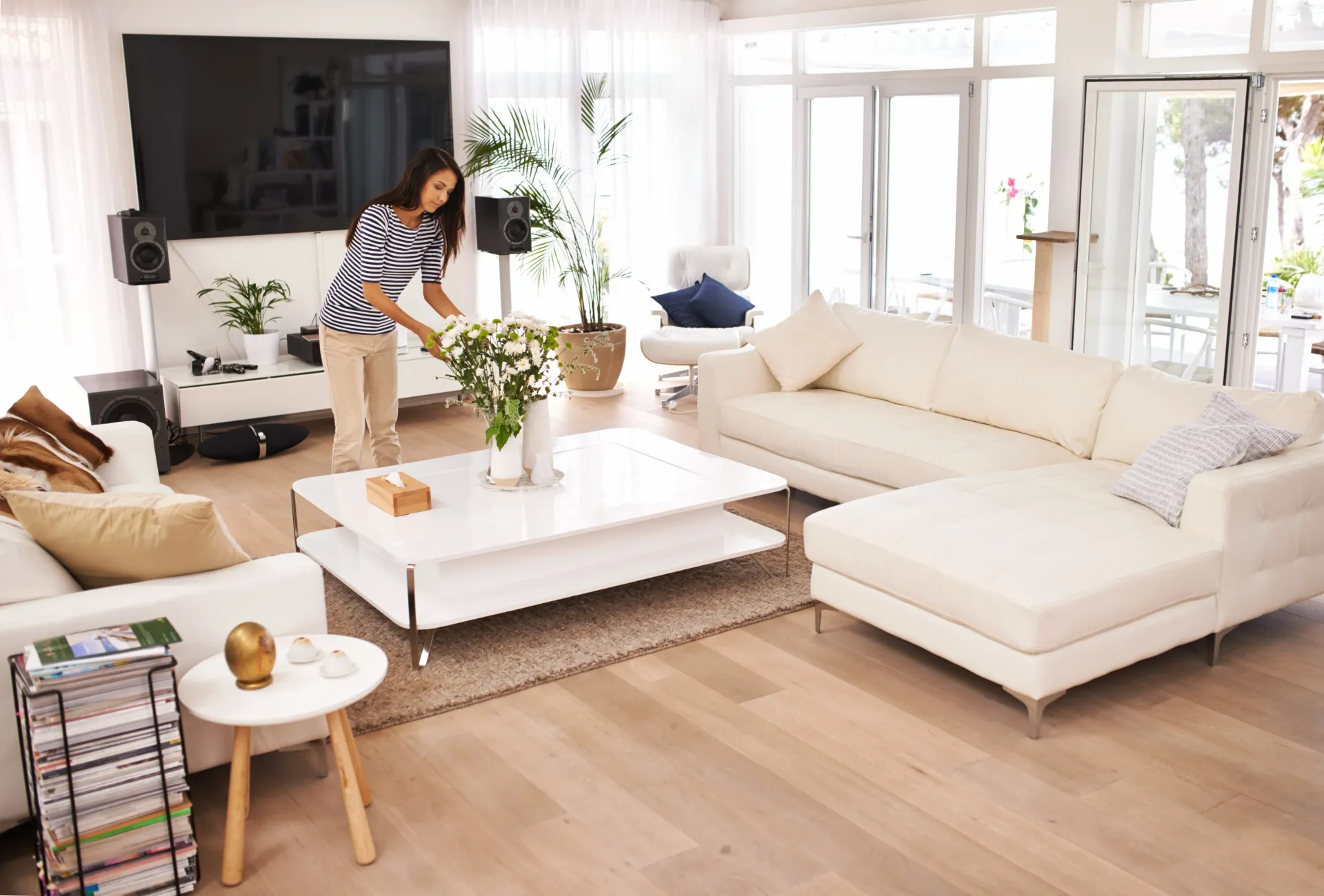 Bright living room with white sectional sofa, coffee table, and a person arranging a plant near a TV.