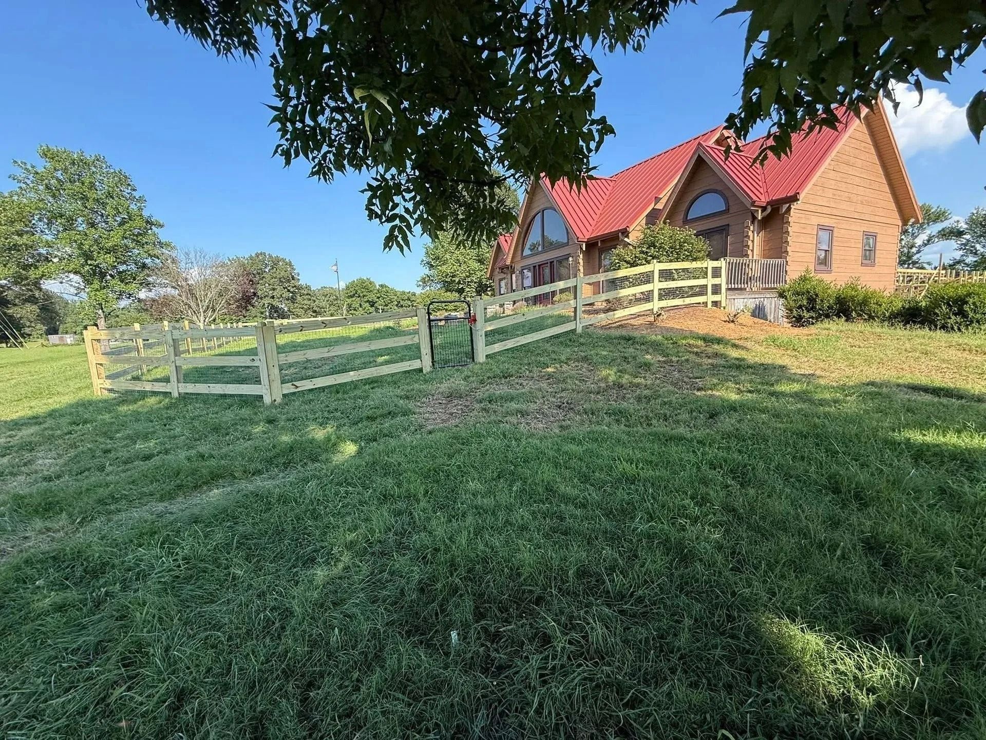 Wooden fence surrounds grassy area, with a brick house and red roof in the background. Blue sky overhead.
