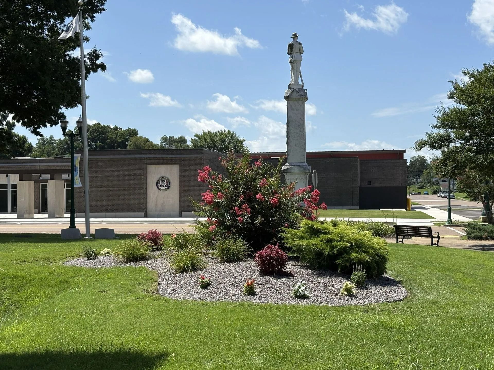 A park with a monument, flowering bushes, and a brick building under a partly cloudy sky.