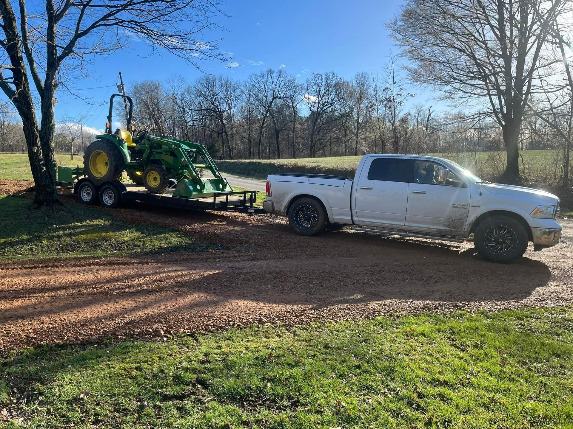 White pickup truck towing a trailer with a green tractor on a gravel driveway in a rural setting.