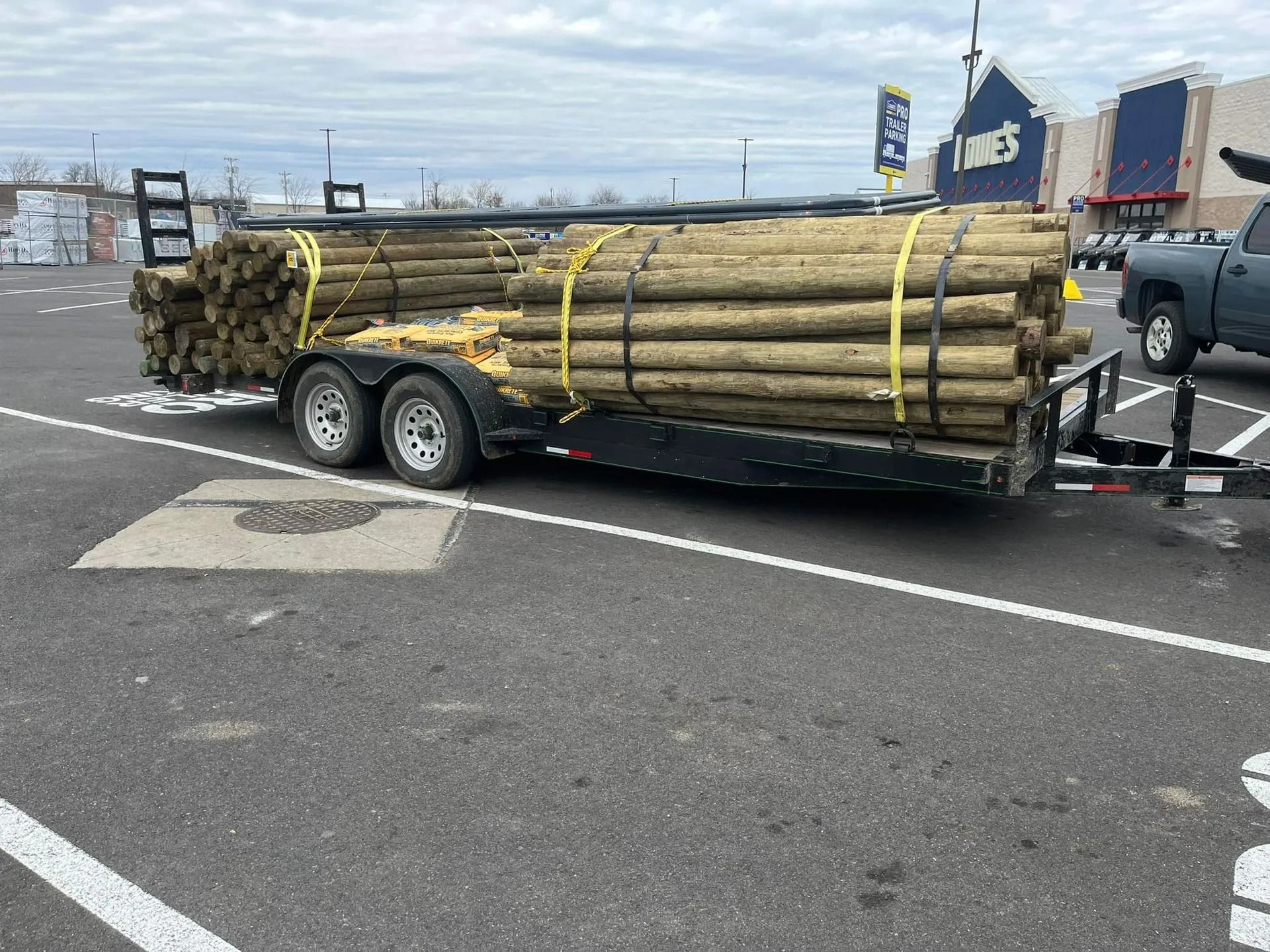 Trailer loaded with wooden fence posts in a Lowe's parking lot. Yellow straps secure the posts.