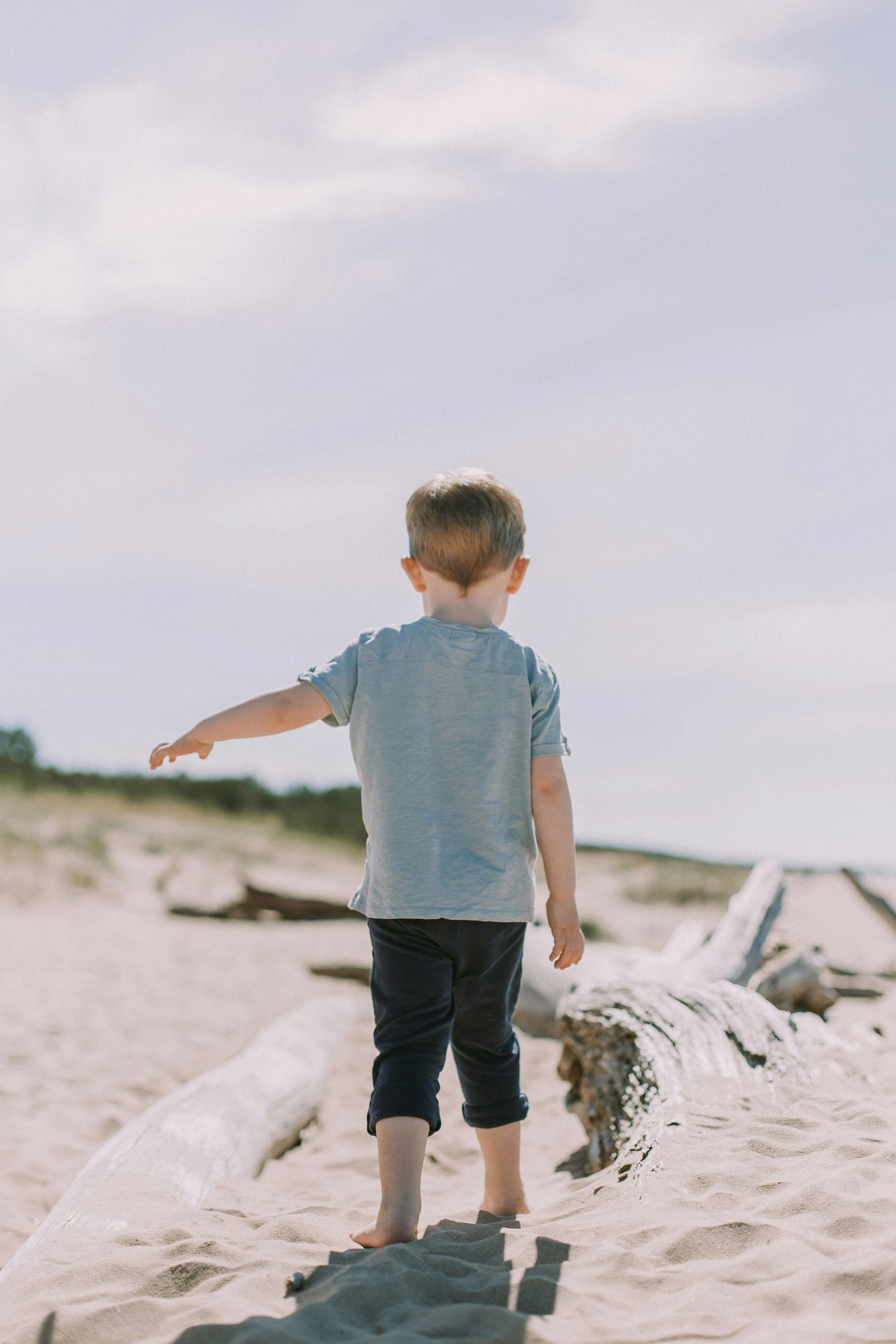 A young boy is standing on a log on the beach.