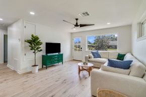 Bright living room with white sofa, teal TV console, wood floor, ceiling fan, and large windows overlooking trees