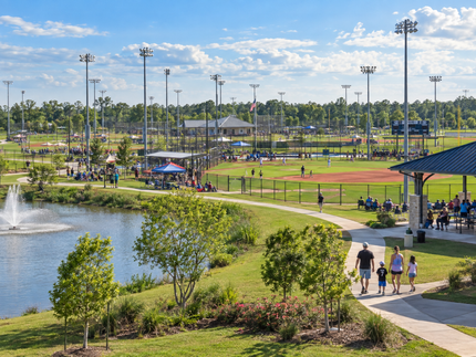 People walking beside a pond and fountain at a sunny baseball park with fields and lights