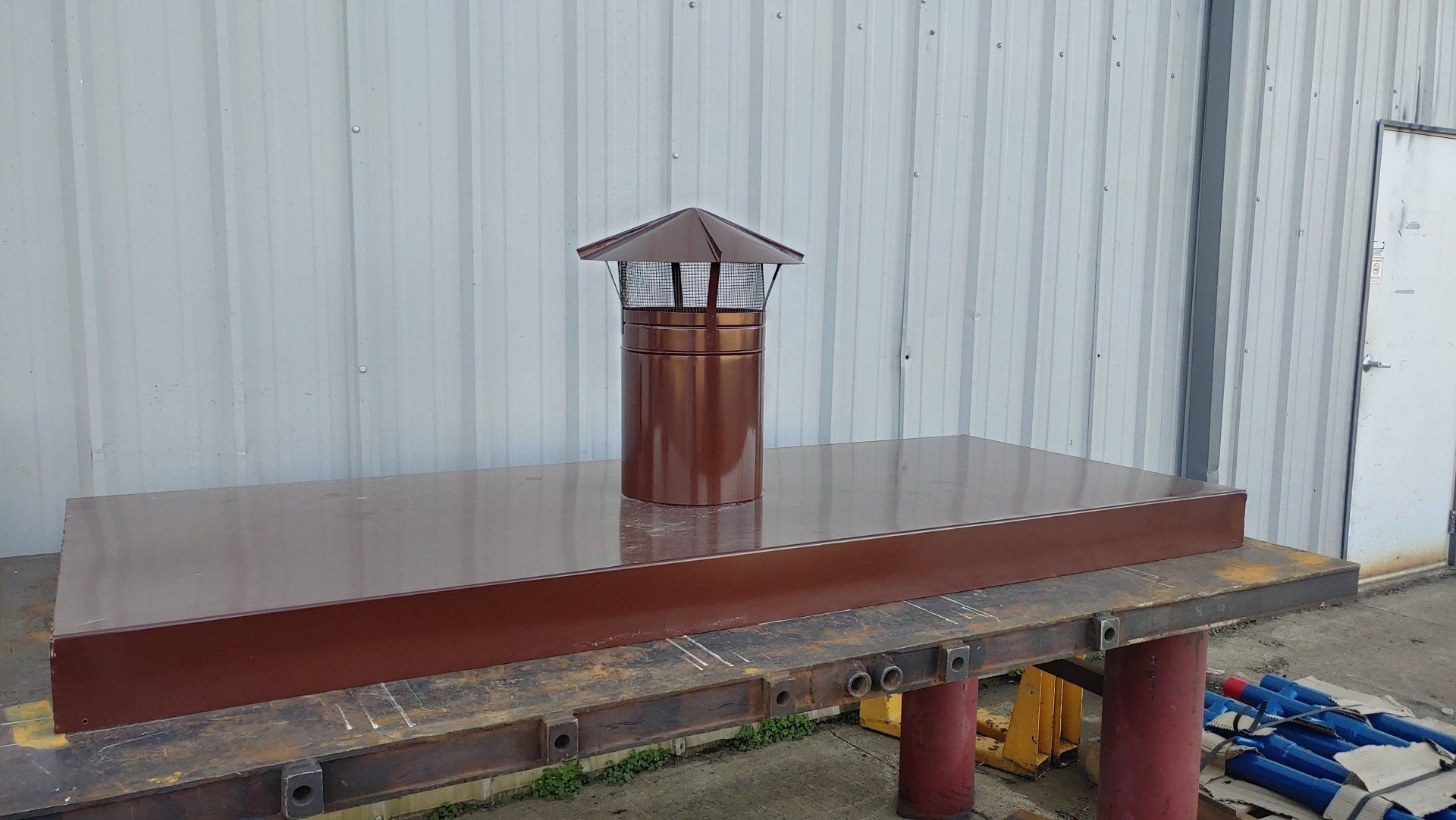 Brown metal chimney on a rectangular base, set on a workbench outdoors.