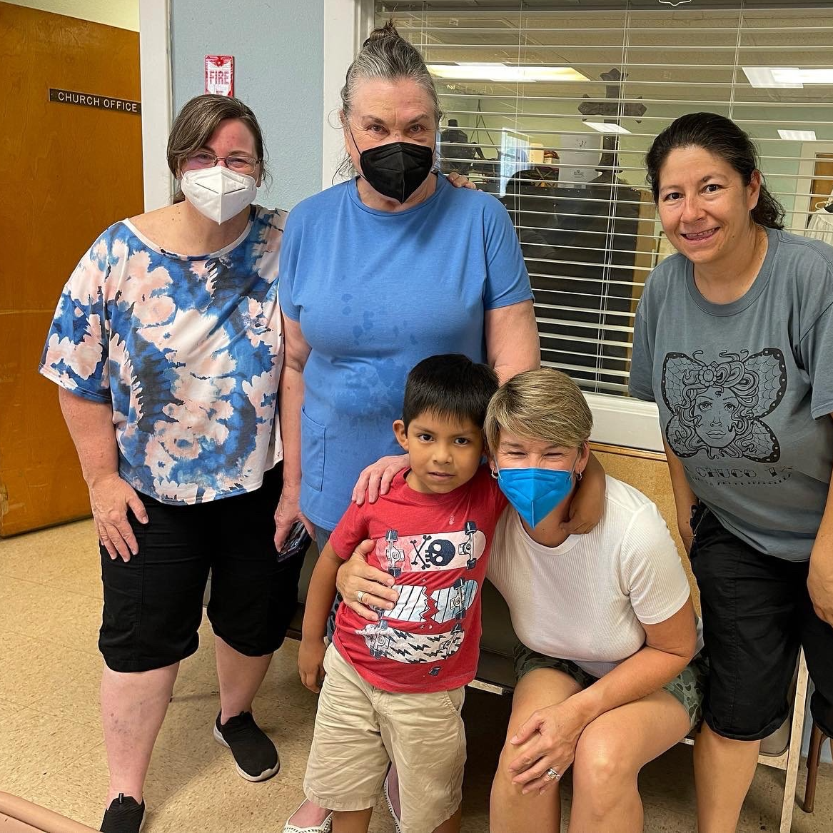 Five masked people pose together indoors, two crouching in front, in a hallway or lobby.
