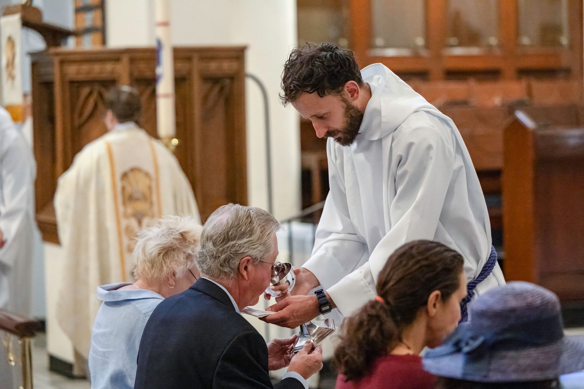Person in white robe giving communion to seated worshippers in a church