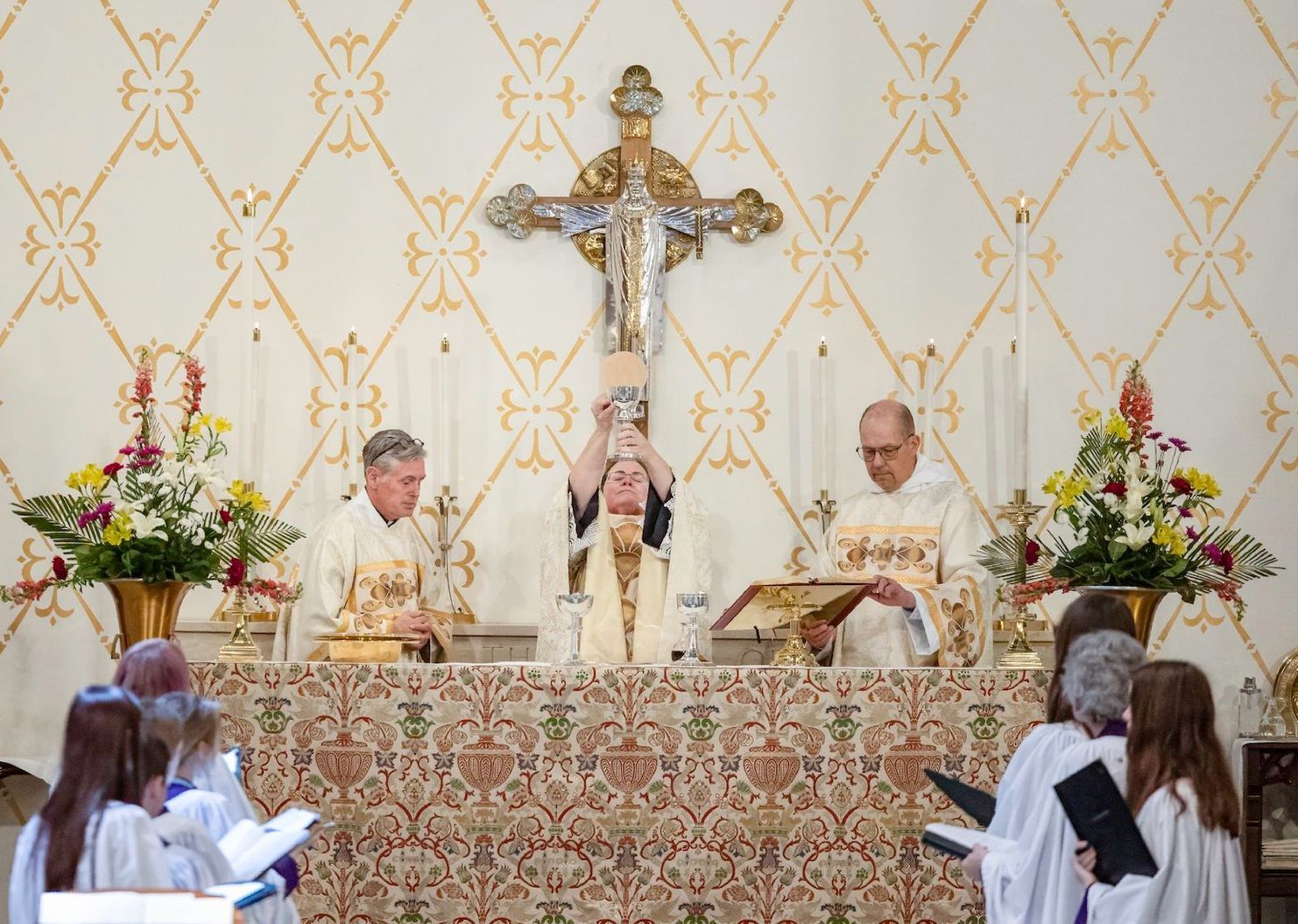 Priest celebrating Mass at an altar with crucifix and floral arrangements, congregation seated in front
