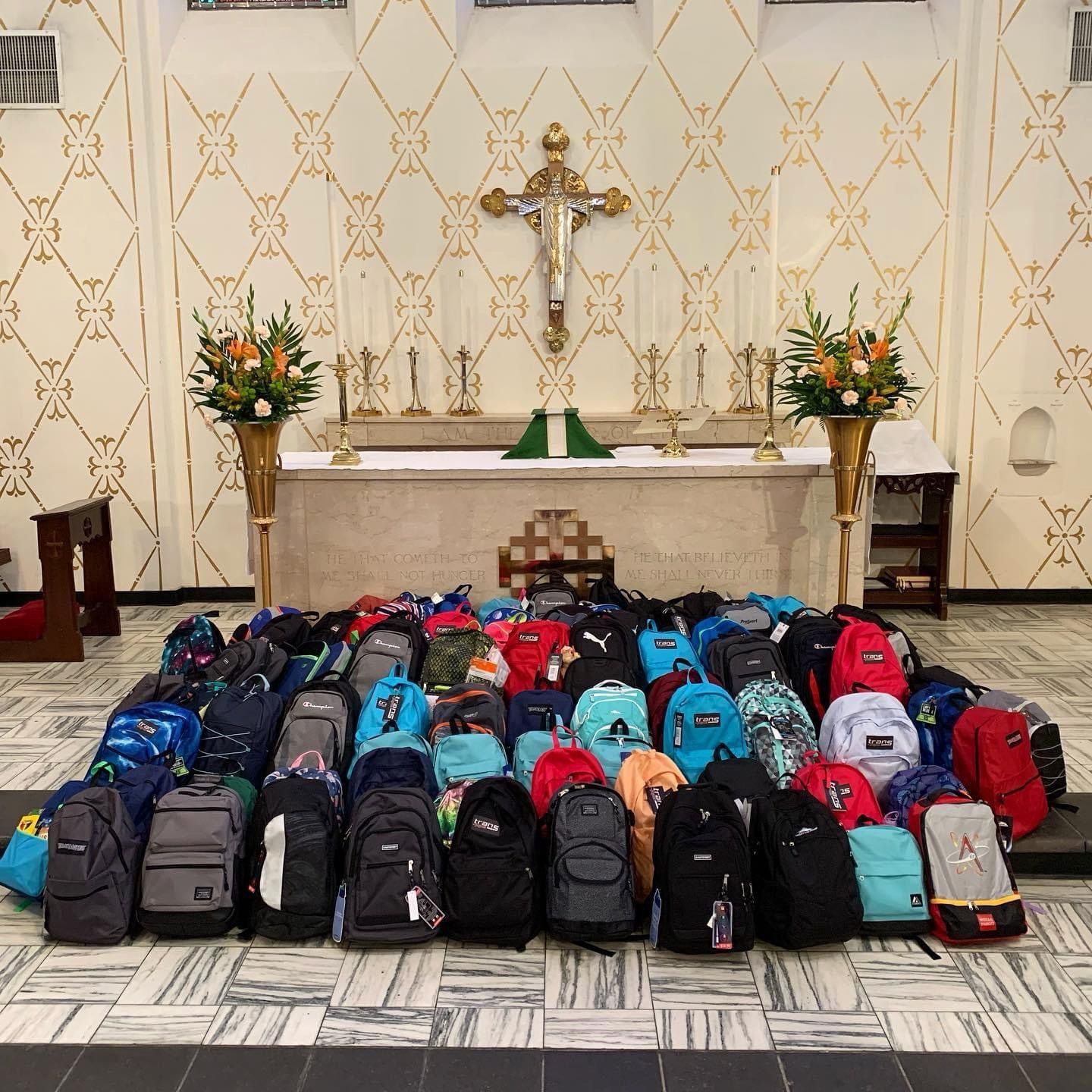 Rows of backpacks arranged before a church altar with a crucifix and flower arrangements for Cathedral of St John's Backpack drive