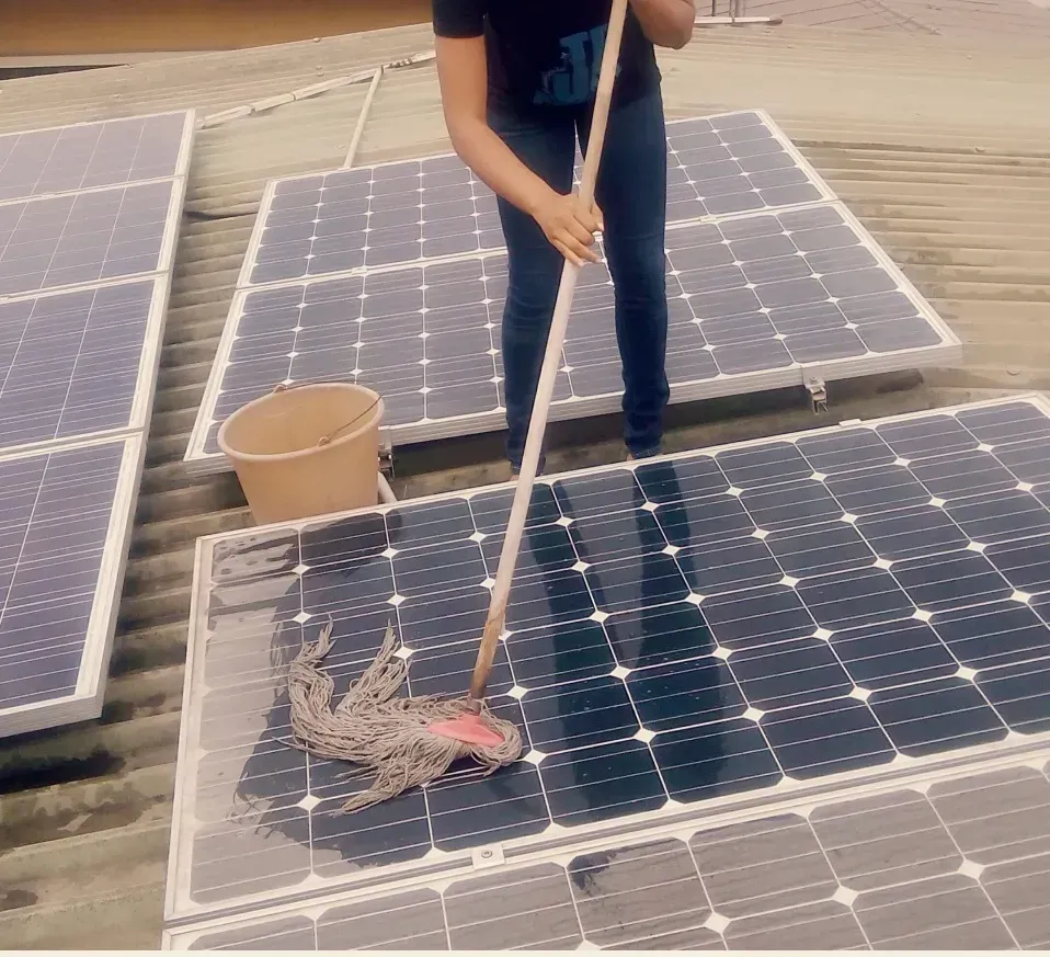 Person cleaning solar panels with a mop on a rooftop, with panels and a bucket nearby.