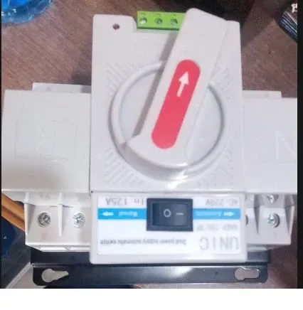 White electrical switch box with a large gray rotary handle and red arrow, mounted on a metal base.