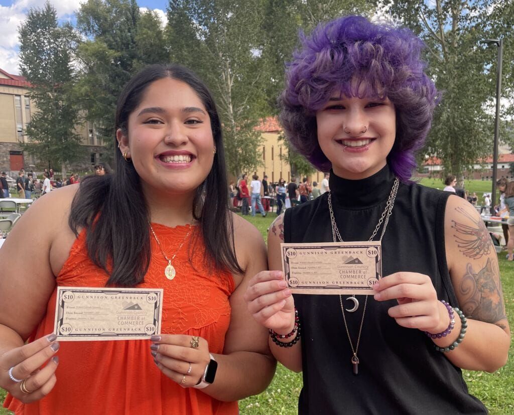 Two people smiling, holding tickets. One in an orange top, the other with purple hair and a black top. Outdoors.