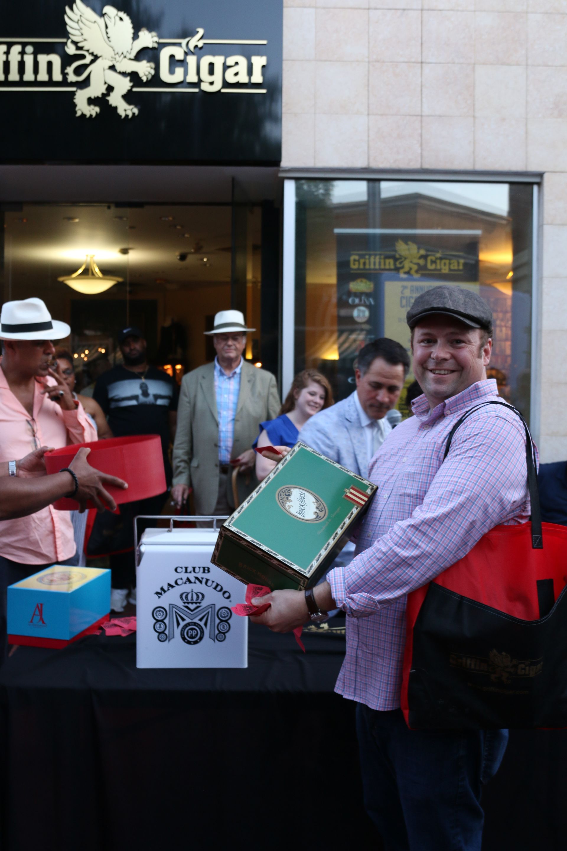 Man holding a green cigar box outside a cigar shop, with people and tables in the background