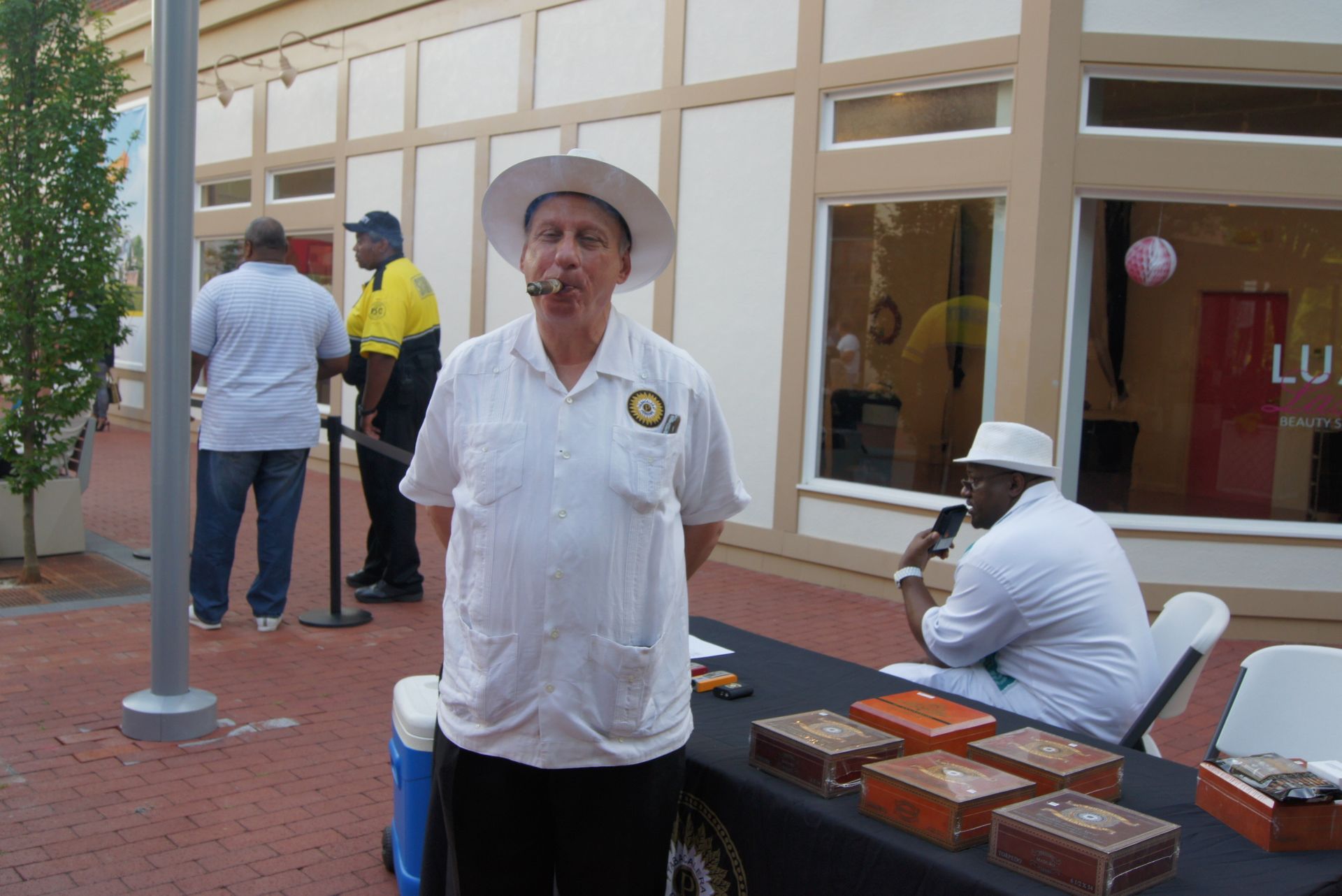 Man in white shirt and hat stands outdoors near a table with pastries, beside seated people and storefront windows