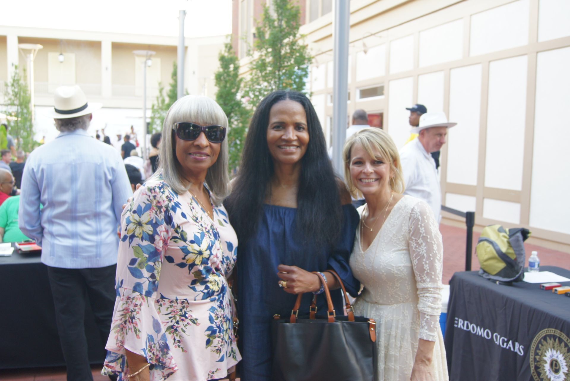 Three women smiling at an outdoor event, standing beside a table with a black cloth.