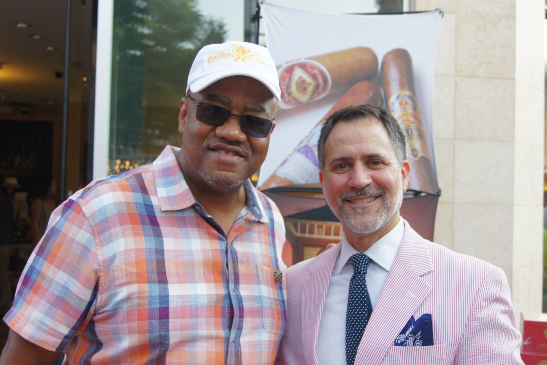 Two men smiling outdoors, one in a plaid shirt and cap, the other in a pink blazer and tie.