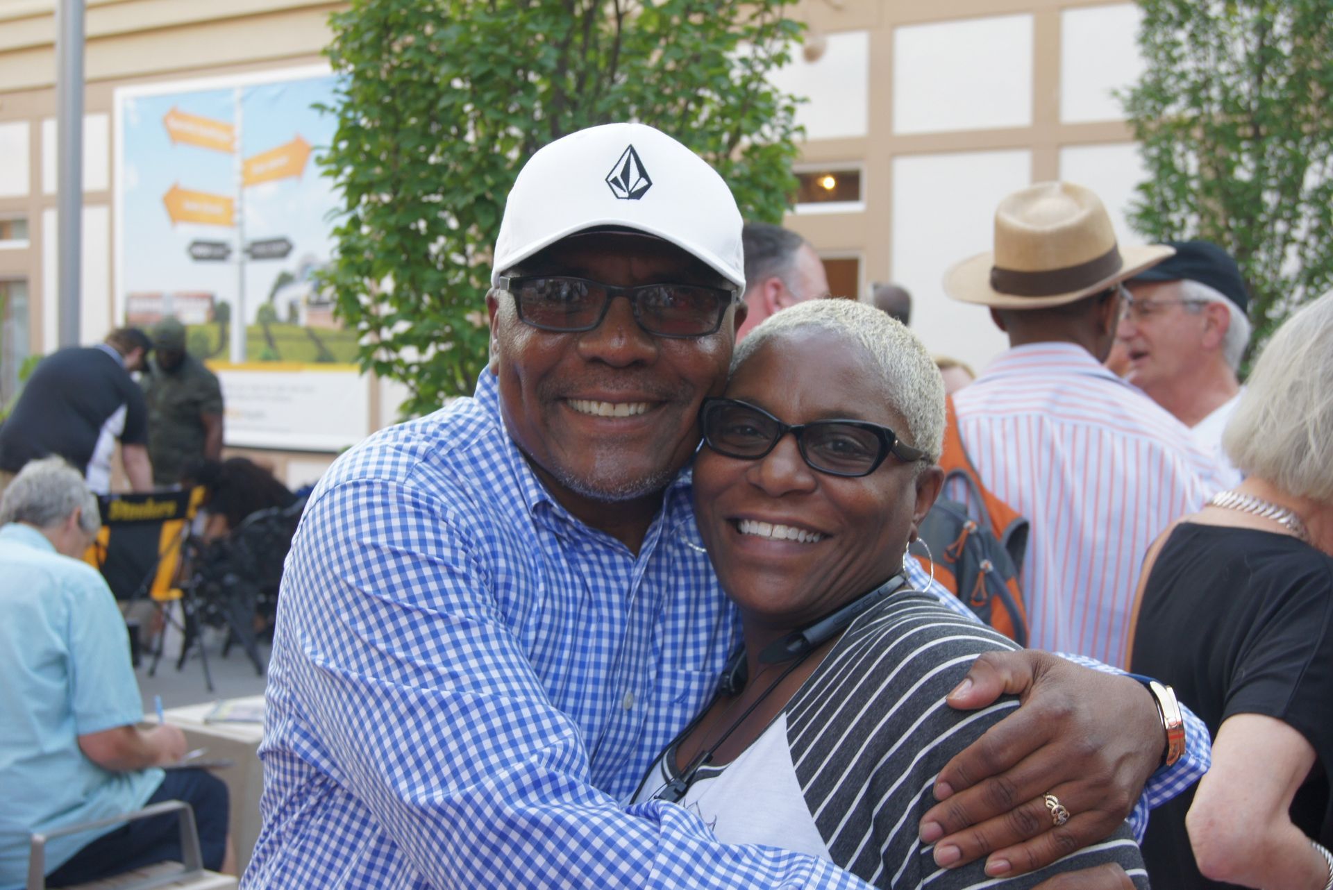 Two smiling people hugging outdoors at a lively event, with other attendees and trees in the background.