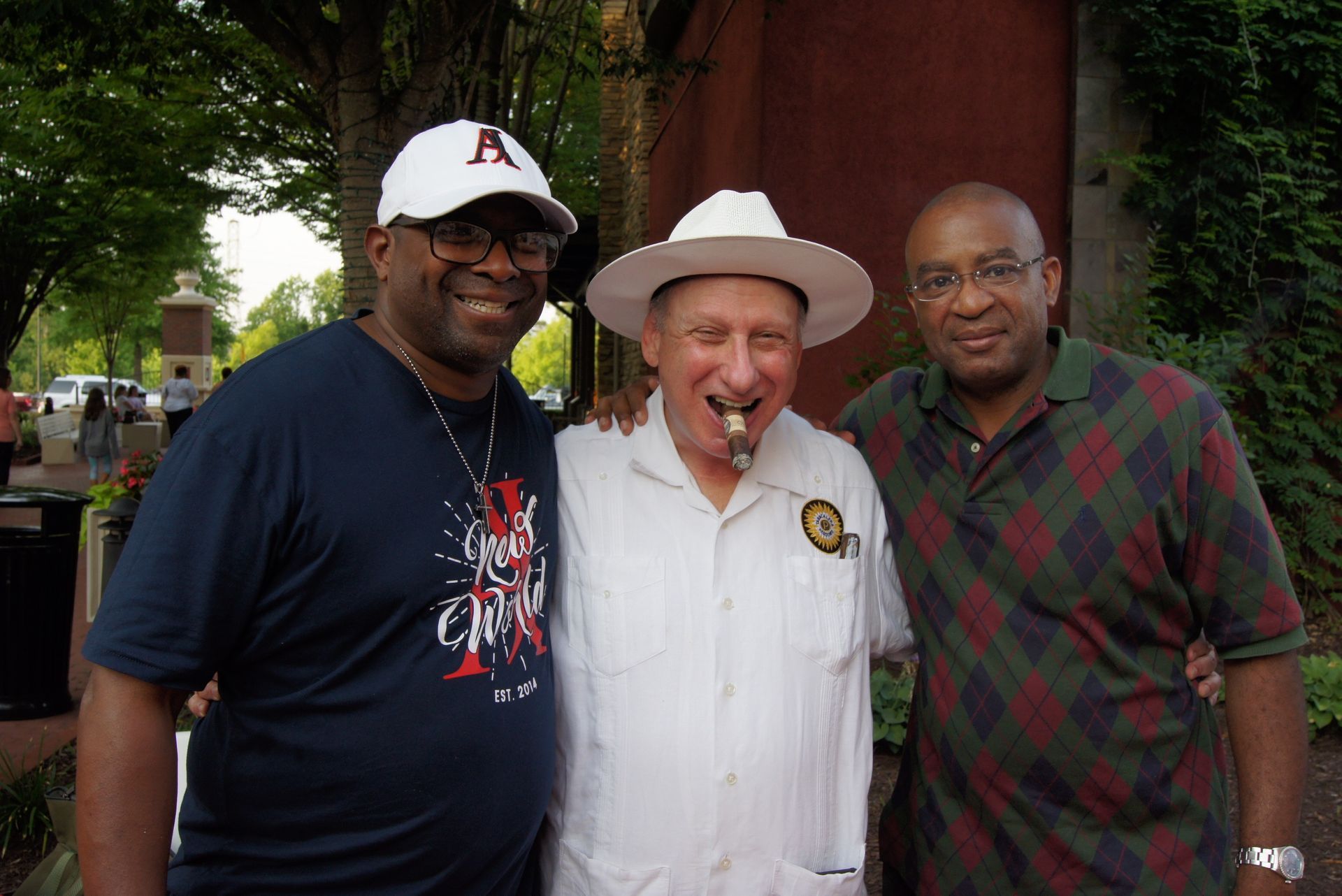Three men pose outdoors, with the center man wearing a white hat and white shirt, smiling.
