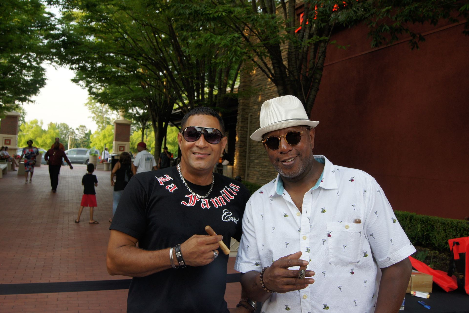 Two men standing on a tree-lined walkway, smiling and holding drinks.