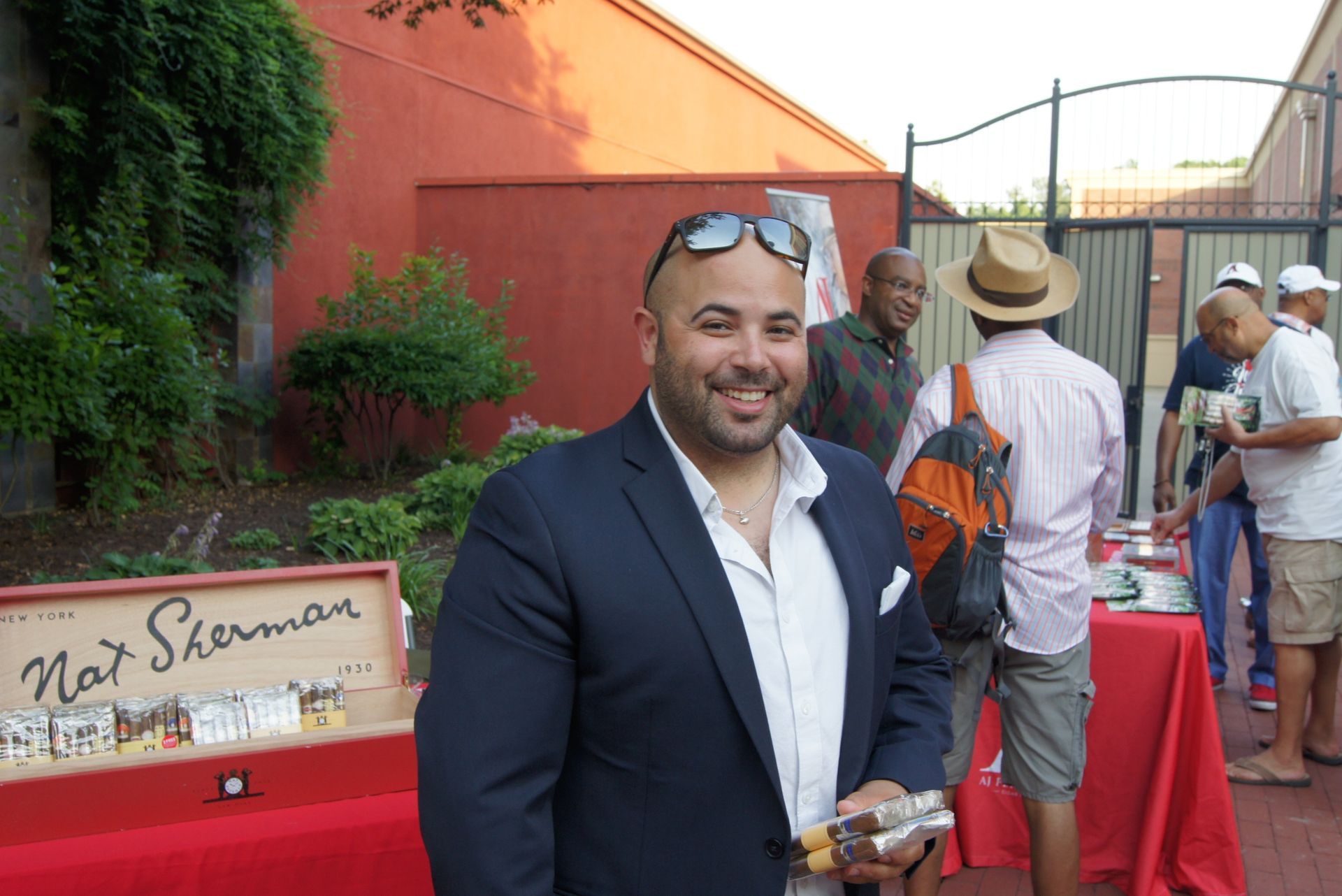 Smiling man in navy blazer at an outdoor event beside red cocktail tables and a gate