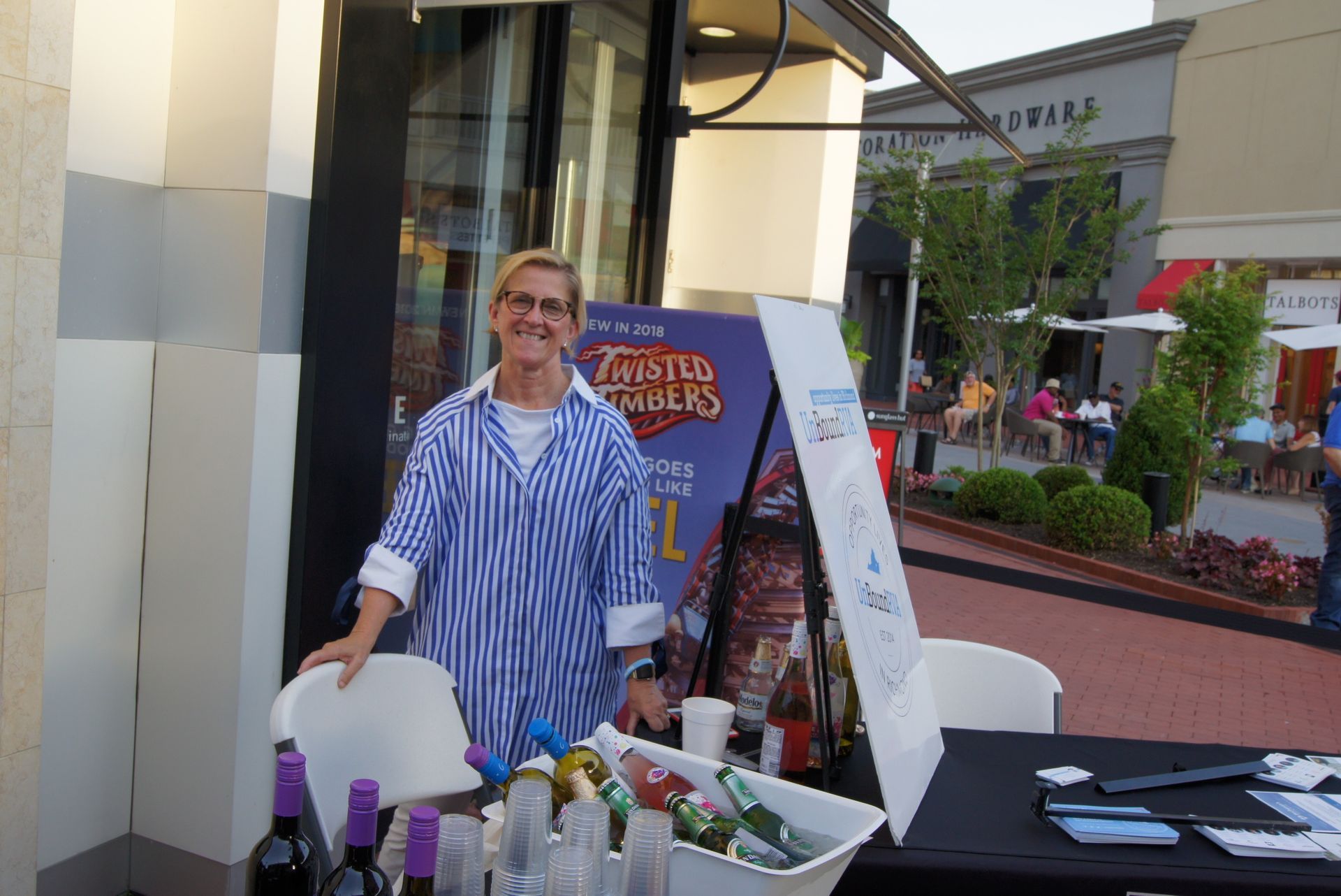 Woman at an outdoor booth with bottled products, a sign, and a table display on a sidewalk.