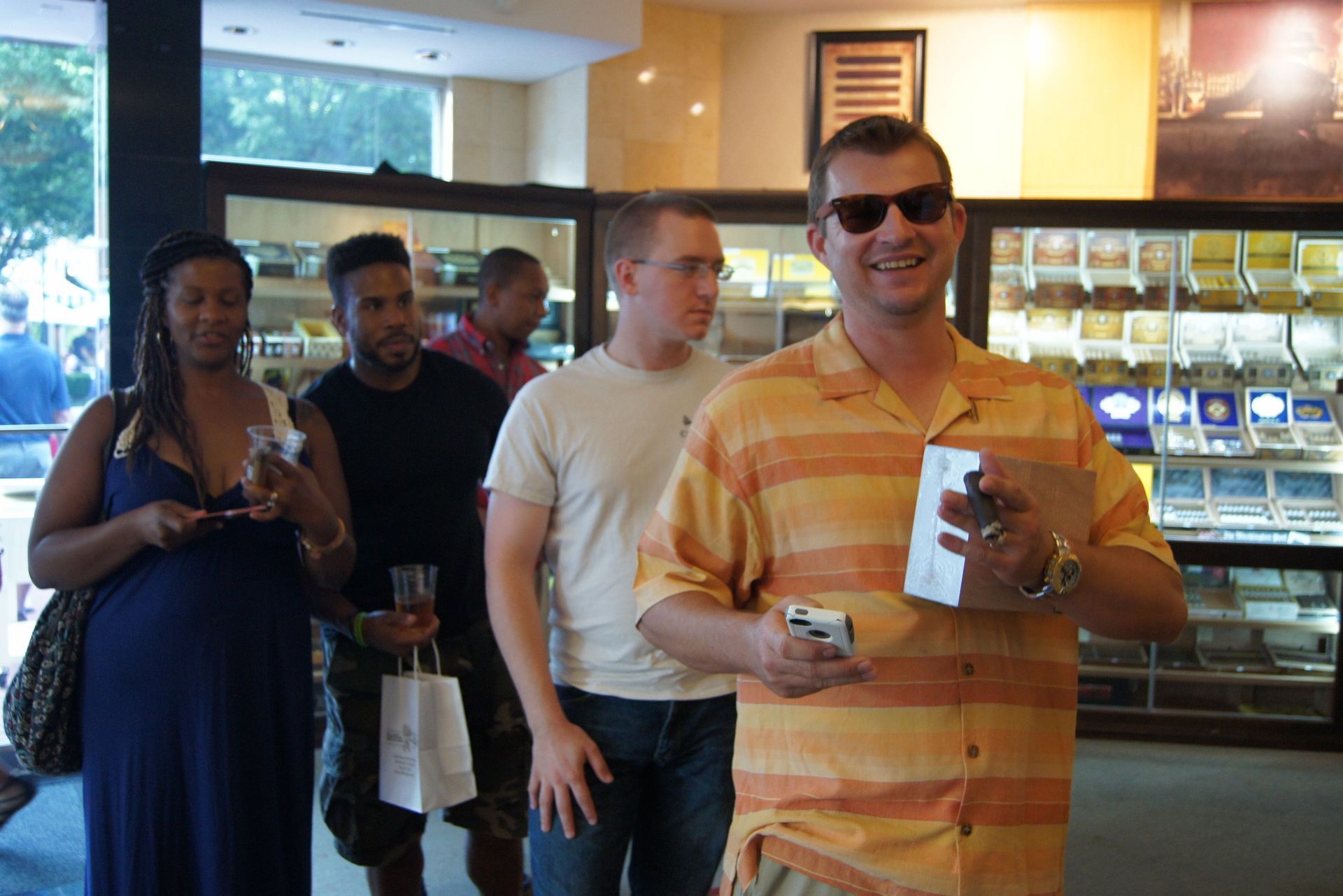 People standing in a café; one man in sunglasses and an orange striped shirt smiles while holding a drink.