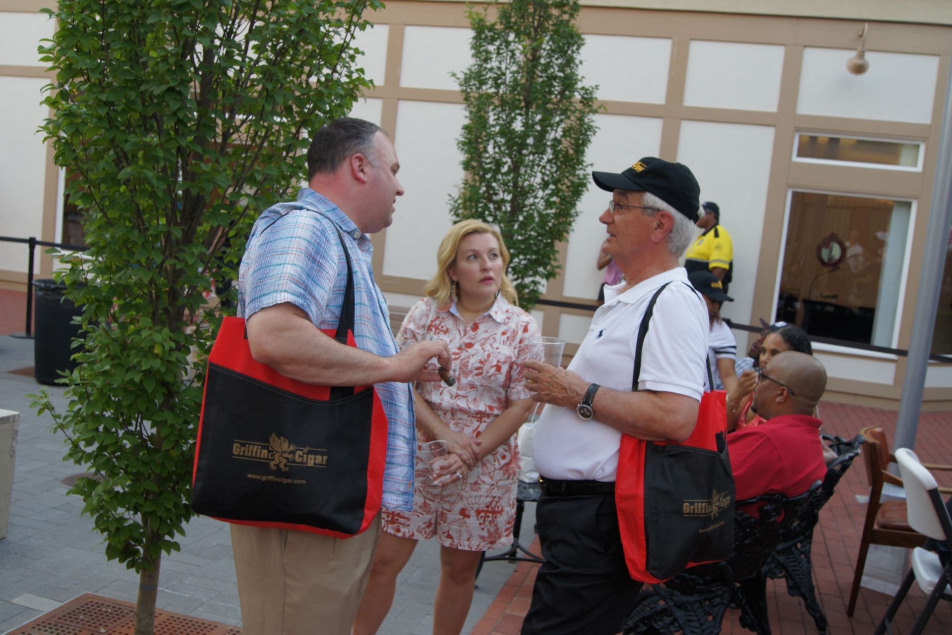 Three people talking outdoors near a building entrance, two holding red-and-black tote bags.