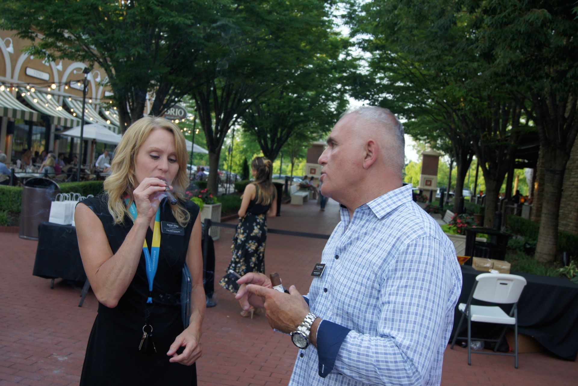 Two people talking outdoors in a tree-lined plaza, one holding a microphone.
