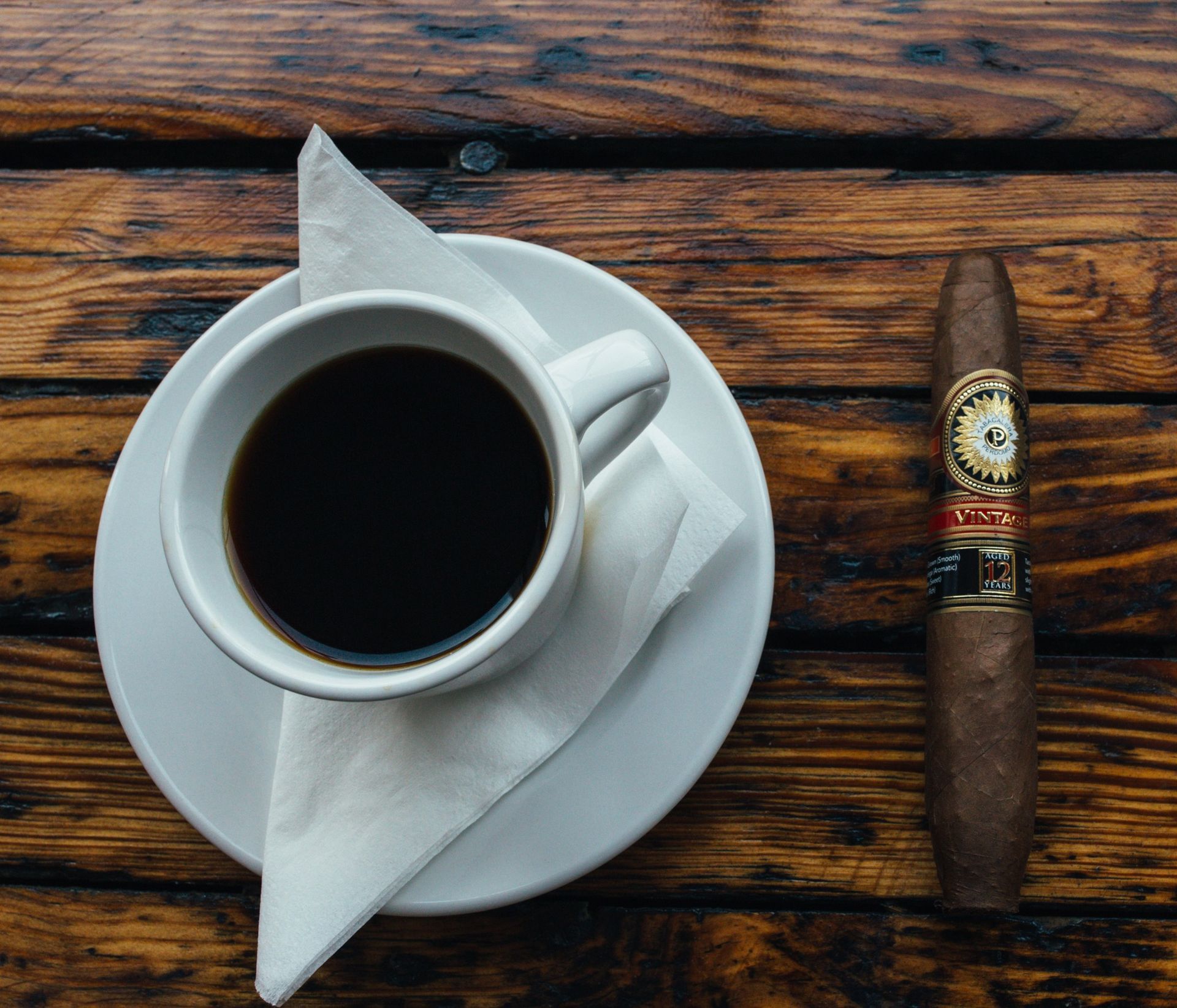 Top-down coffee cup on a saucer with napkin and a cigar on a rustic wooden table.