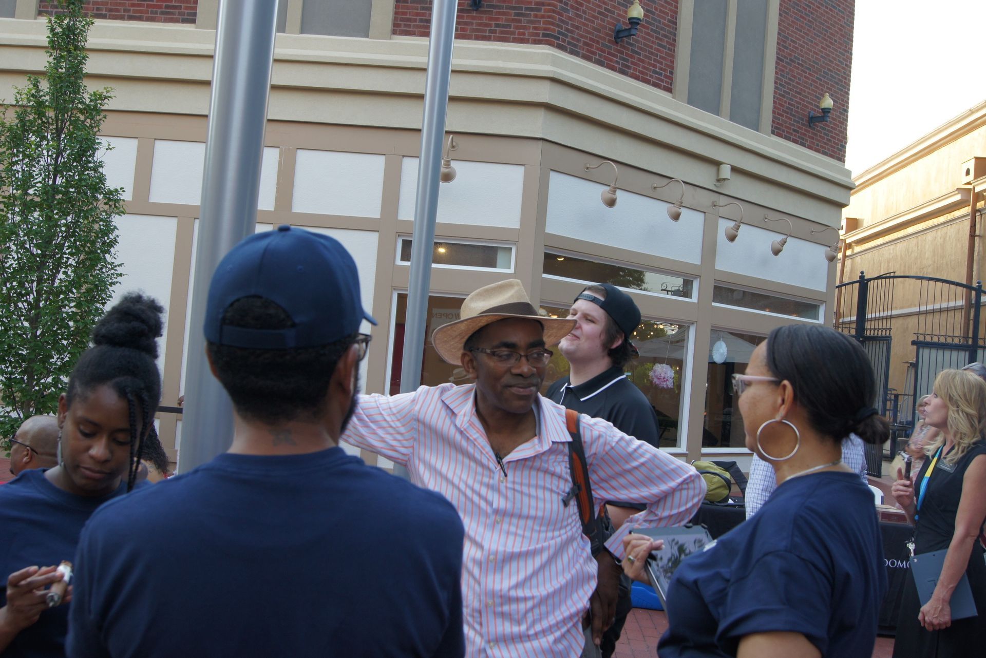 People talking in a courtyard beside a brick building, with a beige awning and trees nearby