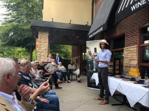 Outdoor wine-tasting talk beside a winery patio with seated audience and speaker in a hat