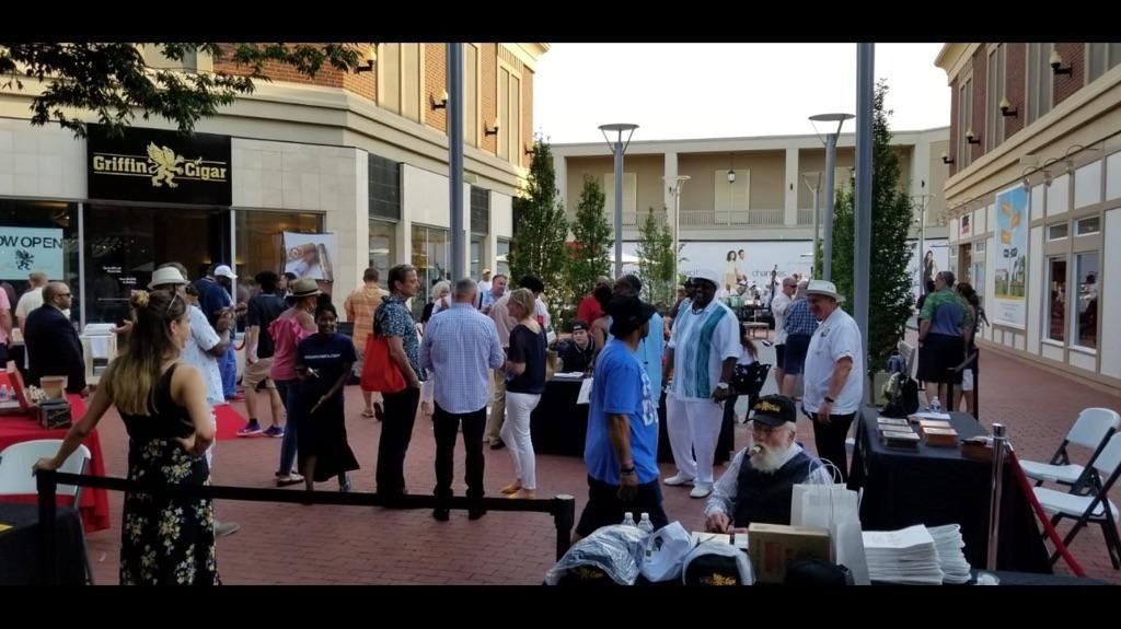 Crowd gathered in a sunny outdoor shopping plaza with shops and tables nearby