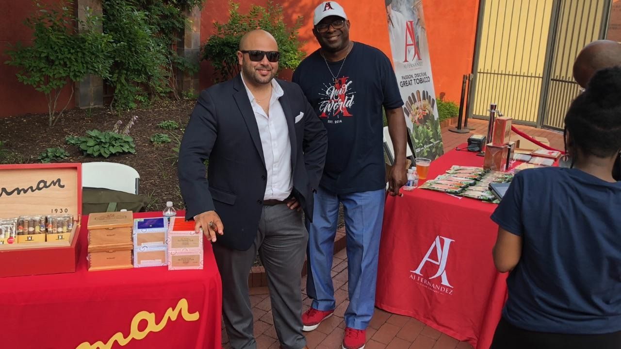 Two men posing at an outdoor event booth with red tablecloths and merchandise display