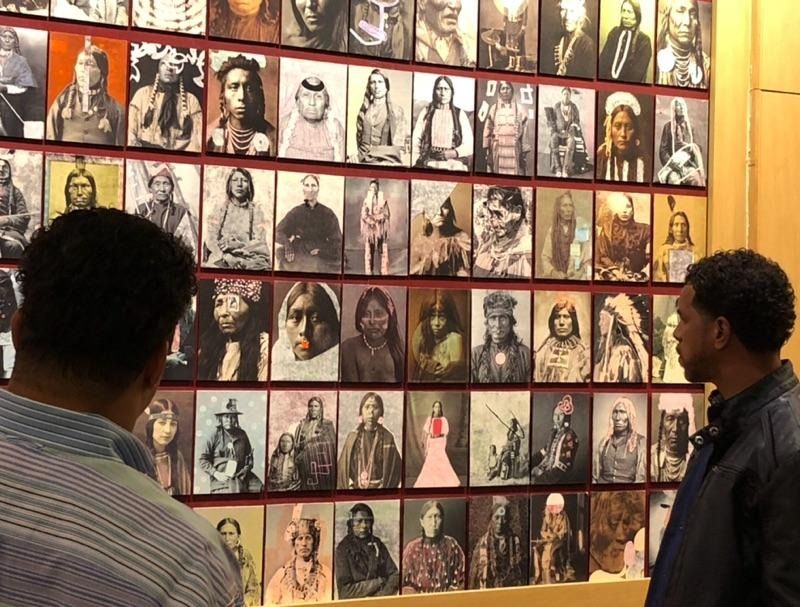 Two people viewing a wall of framed portrait photos in a warm-lit gallery.