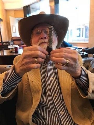 Man in a cowboy hat and vest holding two small objects indoors, seated in a café.
