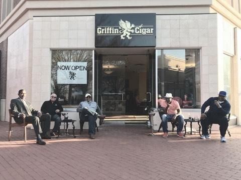 People sitting on benches outside a Coffees and Cigars storefront with a “Now Open” sign.