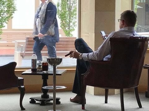 Two men talking in a bright living room, one seated in an armchair and one standing by the window.