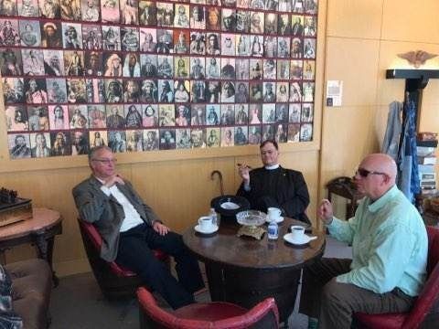 Three men sit at a café table, with cups and saucers in front of a wall covered in photos.