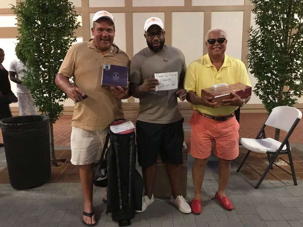 Three men pose indoors holding prize boxes in front of a wall with potted plants.