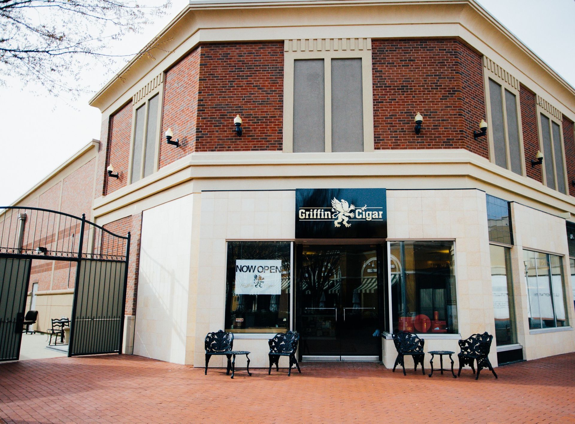 Brick storefront with glass doors and windows, benches outside, and a gated side entrance on a sidewalk.