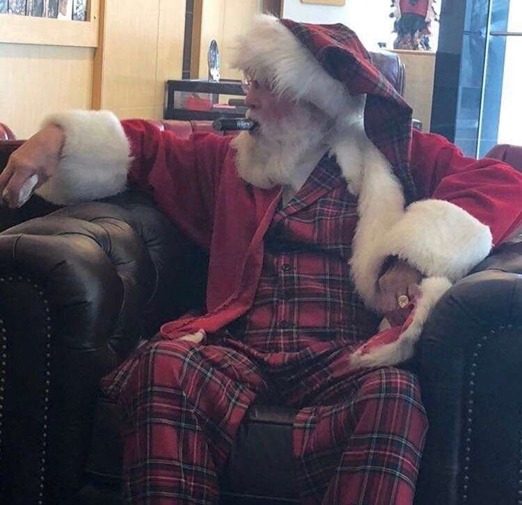 Santa Claus in red plaid sits on a leather chair with a stuffed white cat on his lap.