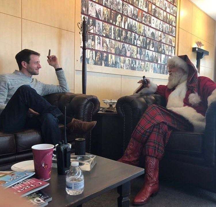 Two people chatting in a lounge, one dressed as Santa in red plaid pants and boots.