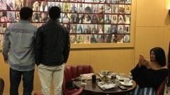 Two people stand facing a wall of photos while a woman sits at a table in a restaurant.