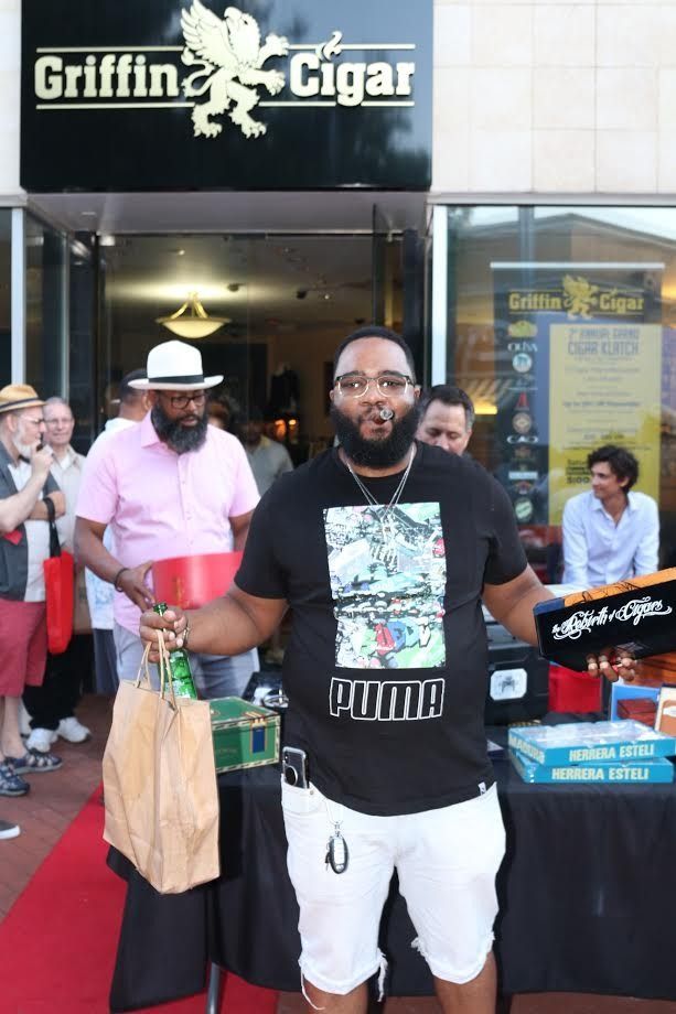 Man standing at a vendor booth with Griffin Cigars sign, holding a bag and wearing a black T-shirt and white shorts