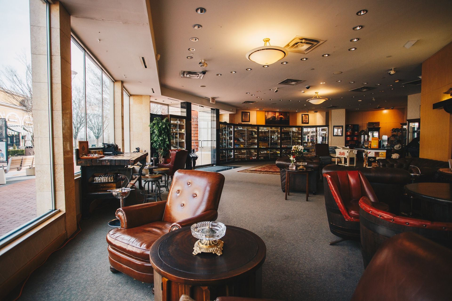Cozy lounge with leather chairs, large windows, warm lighting, and a bar in the background