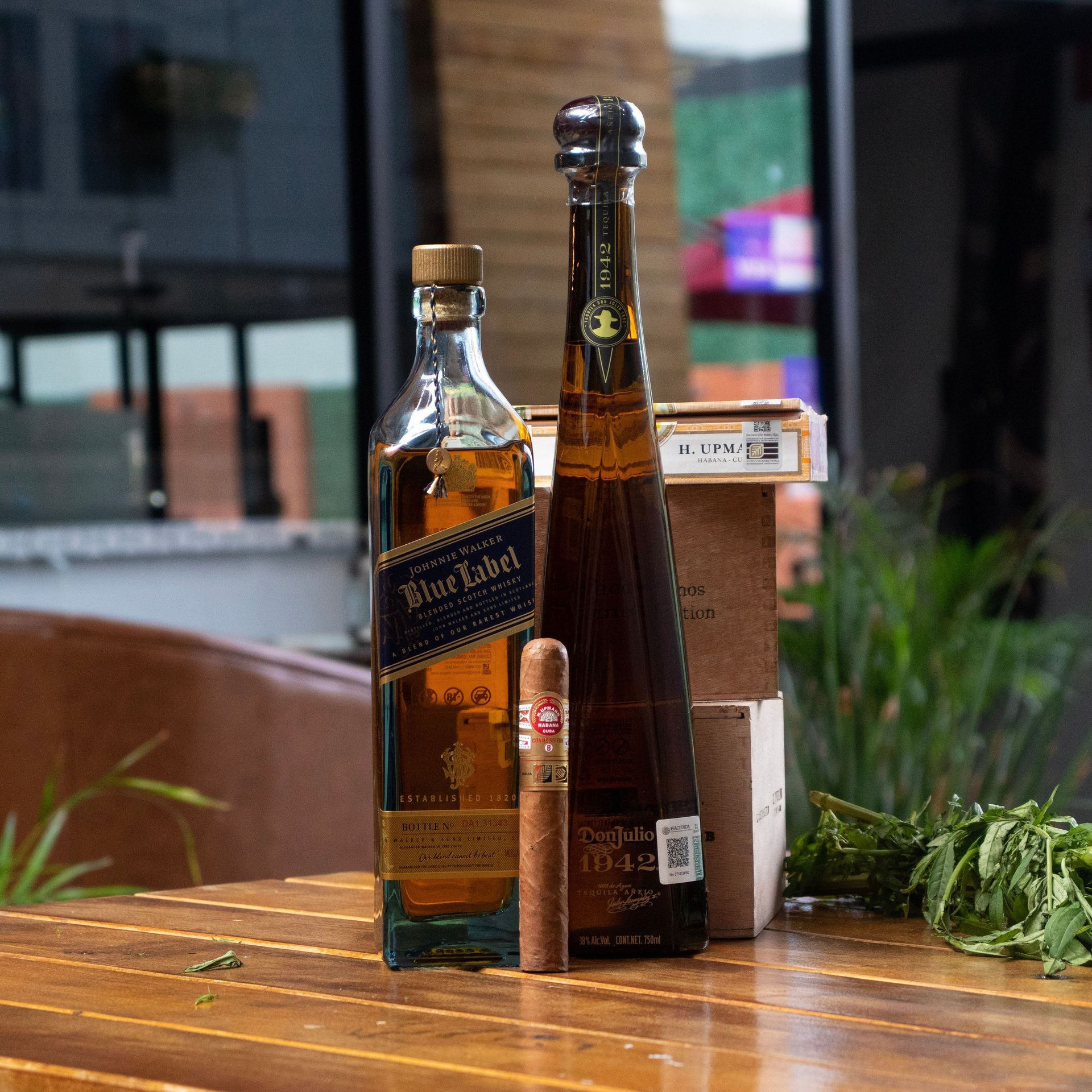 Two liquor bottles and a cigar on a wooden table beside herbs in a café setting