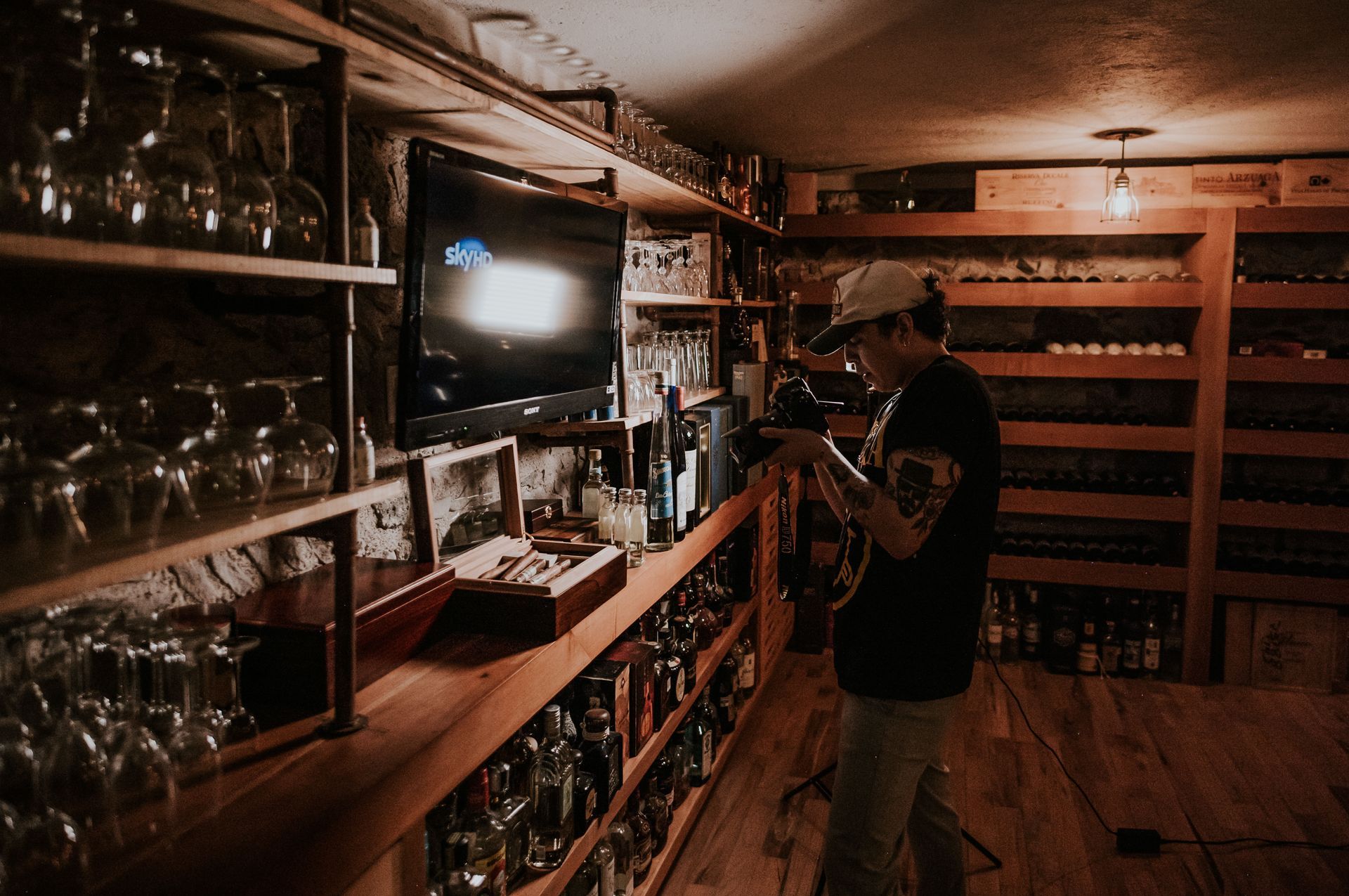 Man in a dim wine cellar arranging bottles on shelves beside glassware and a small TV