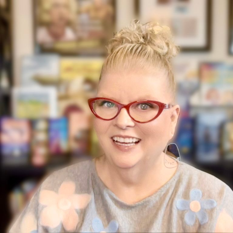 Woman with blonde updo wearing red glasses smiles, gray shirt with blue flowers; books in background.